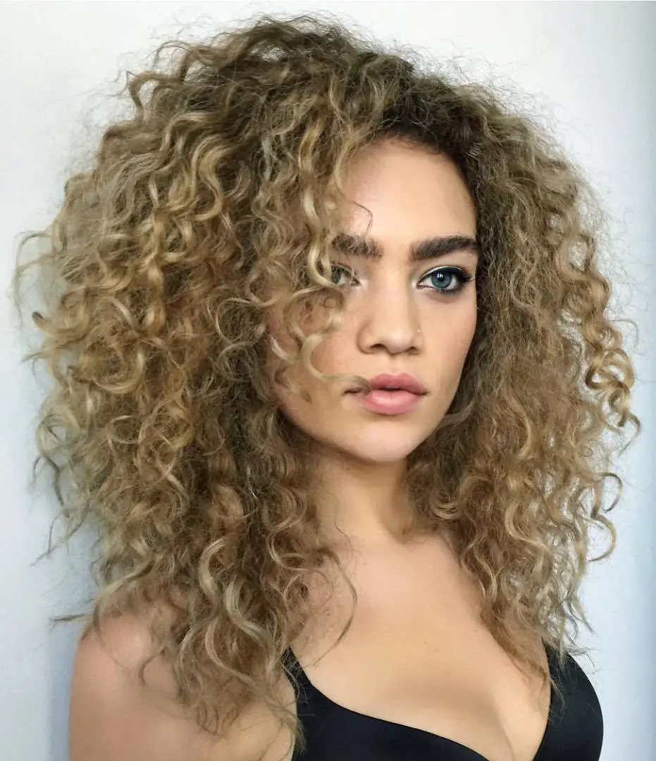 Femme aux cheveux bouclés avec une coupe dégradée volumineuse, racines foncées et sourcils marqués pour un look audacieux et sexsuel.
