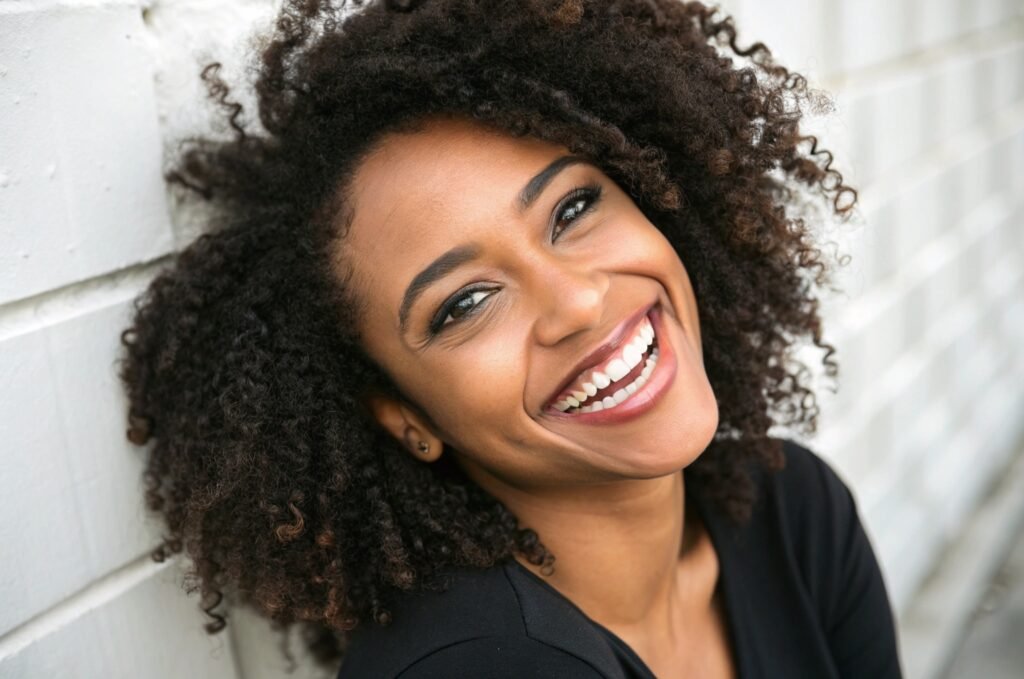 Confiance et Éclat - Personne aux Cheveux Bouclés Rayonnants Personne aux cheveux bouclés rayonnants de confiance, souriant à la caméra, illustrant l'impact positif d'une routine de soins capillaires pour des boucles bien définies et en bonne santé.